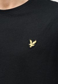 Μαύρο μπλουζάκι με στρογγυλή λαιμόκοψη και μικρό κίτρινο κεντητό λογότυπο πουλιού στο αριστερό στήθος, εμφανίζεται στο άνω μέρος του κορμού ενός ατόμου.