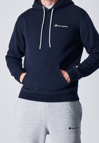 Sudadera de color marino en material suave con bolsillo tipo canguro, cordones blancos y un pequeño logo de Champion bordado en el pecho. Combinada con pantalones de chándal grises.