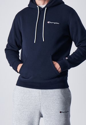Navy Kapuzenpullover aus weichem Material mit einer Kängurutasche, weißen Kordeln und einem kleinen, auf die Brust gestickten Champion-Logo. Kombiniert mit grauen Jogginghosen.