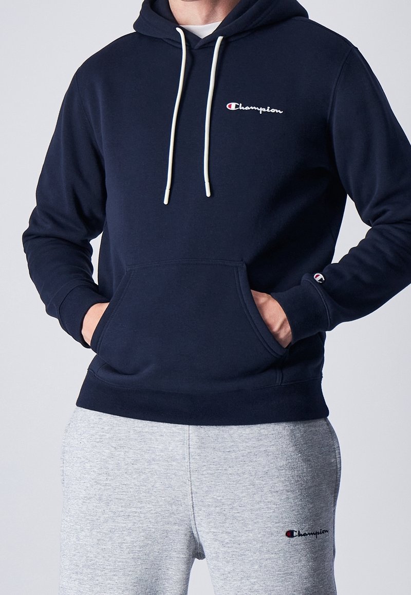 Sudadera de color marino en material suave con bolsillo tipo canguro, cordones blancos y un pequeño logo de Champion bordado en el pecho. Combinada con pantalones de chándal grises.