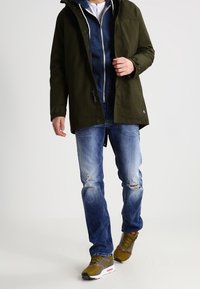 Chaqueta impermeable verde oliva con capucha sobre una sudadera con capucha azul marino, vaqueros desgastados azul claro y zapatillas verde oliva con detalles rojos.