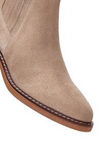 Stivale alla caviglia in suede beige con punta appuntita, suola marrone a contrasto e sottili dettagli di cucitura lungo il bordo. Tessuto liscio nel complesso.