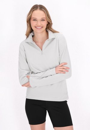 Lys grå fleece-pullover med kvart lynlås, lange ærmer og blød tekstur, parret med sorte shorts, model stående med armene korslagt.