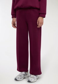 Pantaloni sportivi color marrone scuro con vestibilità ampia, tessuto strutturato, vita elasticizzata e tasche laterali, abbinati a scarpe da corsa argento.