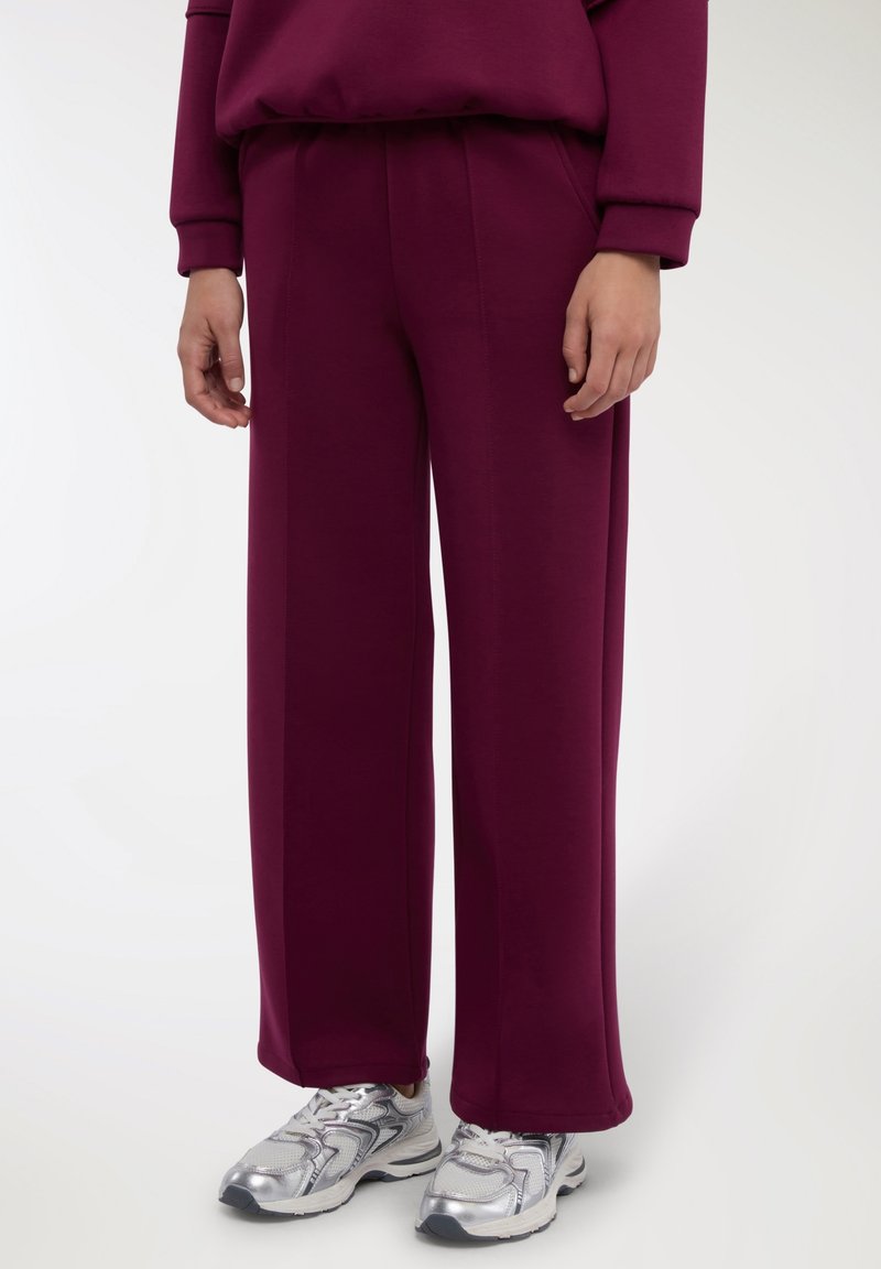 Pantaloni sportivi color marrone scuro con vestibilità ampia, tessuto strutturato, vita elasticizzata e tasche laterali, abbinati a scarpe da corsa argento.