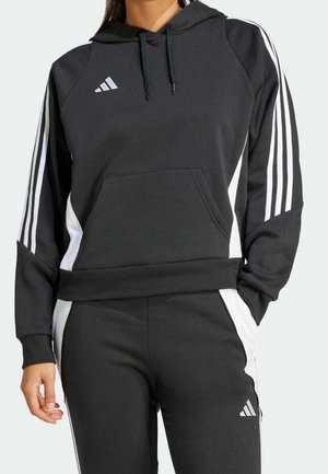 Frau trägt schwarzen Adidas Hoodie und passende schwarze Jogginghose mit weißen Streifen und Logo an Ärmeln und Beinen.