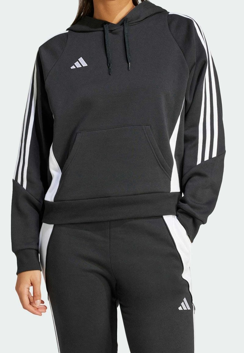 Femme portant un sweat à capuche noir Adidas et un pantalon de survêtement noir assorti avec des bandes blanches et un logo sur les manches et les jambes.