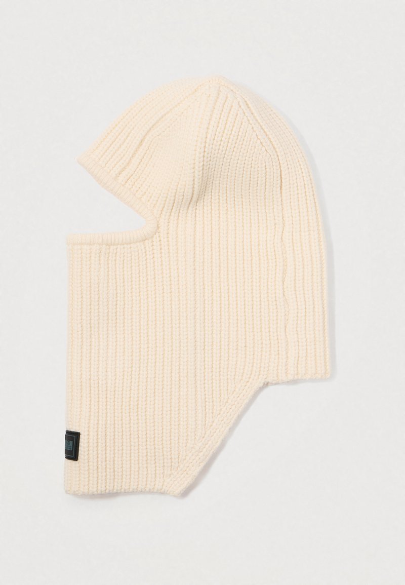 Balaclava en tricot beige avec texture côtelée, design couvrant intégralement et découpes latérales pour le visage. Dotée d'une petite étiquette noire au bas.