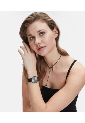 La montre présente un cadran rond argenté-gris avec des index horaires noirs et un bracelet en cuir noir texturé. Portée au poignet d'une femme.