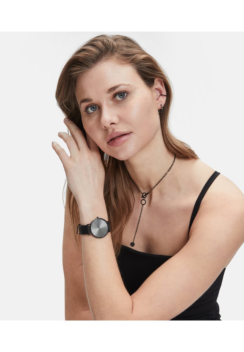 Die Uhr verfügt über ein rundes, silbergraues Zifferblatt mit schwarzen Stundenmarkierungen und einem strukturierten schwarzen Lederarmband. Sie wird am Handgelenk einer Frau getragen.