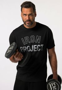 Camiseta de manga corta negra con "IRON PROJECT" en letras gris oscuro en negrita, sostenida por una persona que agarra un dumbbell negro con un agarre circular.