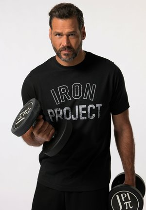 Schwarzes T-Shirt mit kurzen Ärmeln, auf dem "IRON PROJECT" in fetten grauen Buchstaben steht, gehalten von einer Person, die ein schwarzes Hantel mit einem runden Griff greift.