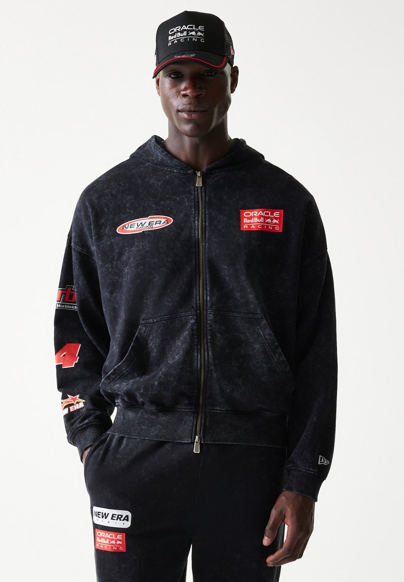 Μαύρη φούτερ ζακέτα με φερμουάρ και ελαφρώς φθαρμένη υφή. Διαθέτει λογότυπα: "Oracle Red Bull Racing" και "New Era." Σκούρο παντελόνι ολοκληρώνει την εμφάνιση.