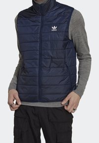 Chaleco acolchado azul marino con cuello alto, cierre de cremallera y bolsillos laterales. Presenta el logo de Adidas en blanco. Llevado sobre una camiseta de manga larga gris.