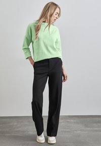 Vaaleanvihreä teksturoitu neulepaita, jossa on korkea kaulus, yhdistettynä muotonsa mukaisiin mustiin housuihin ja valkoisiin jalkineisiin, harmaata taustaa vasten.