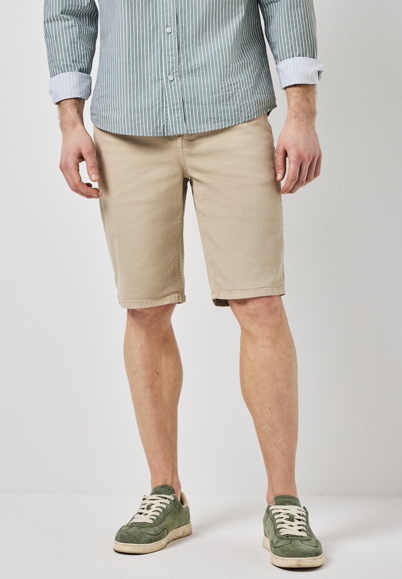 Béžové kraťasy vyrobené z lehké látky, dlouhé po kolena s jemnou texturou. Na sobě mají zelené semišové tenisky a světle pruhovanou košili.