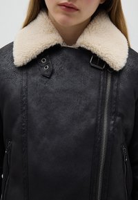 Giacca nera in ecopelle con colletto crema e morbido. Presenta una cerniera laterale, dettagli con bottoni e una finitura testurizzata. Vista ravvicinata.