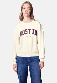 Kremowy sweatshirt z nadrukiem "BOSTON" i "1980" w kolorze bordowym. Posiada okrągły dekolt i ściągacze przy mankietach. W połączeniu z niebieskimi dżinsami.