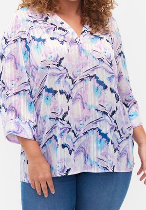 Krøllet person iført en løs, langærmet bluse med lilla, blå og sorte abstrakte mønstre, kombineret med blå jeans.