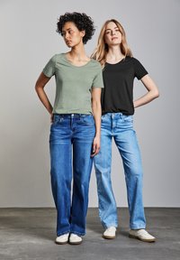 Dos mujeres están de pie una al lado de la otra, vistiendo una camiseta verde oliva y una camiseta negra, combinadas con jeans claros y oscuros, respectivamente.
