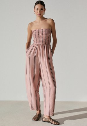 Kobieta w bezramkowym, marszczonym różowym kombinezonie w pionowe paski, stojąca z rękami w kieszeniach i nosząca czarne buty z siateczki.