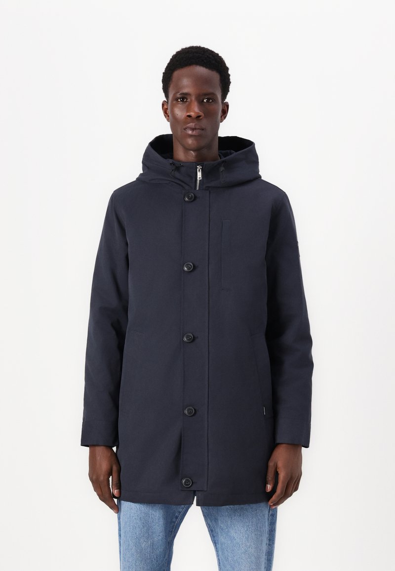 Granatowy płaszcz z kapturem z zamkiem błyskawicznym i zapięciem na guziki, z kieszenią na piersi oraz dwiema kieszeniami po bokach. Wykonany z gładkiego, teksturowanego materiału.
