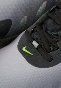 Szare buty sportowe z siateczkową i syntetyczną konstrukcją. Mają czarne sznurówki i zielone logo Nike w kształcie znaku Swoosh. Widoczne są teksturowane sekcje.