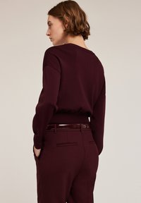 Pulover burgund cu guler rotund și mâneci ușor supradimensionate, purtat cu pantaloni tapered asortați, având buzunare la spate și o centură lucioasă.