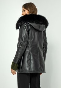 Zwarte leren jas met een grote zwarte bontkap. Beschikt over een.getailleerde snit, een gladde textuur en groene gebreide manchetten. Achteraanzicht weergegeven.