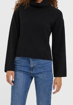 Femme portant un pull à col roulé noir à manches longues et un jean bleu en denim, debout devant un fond clair uni.