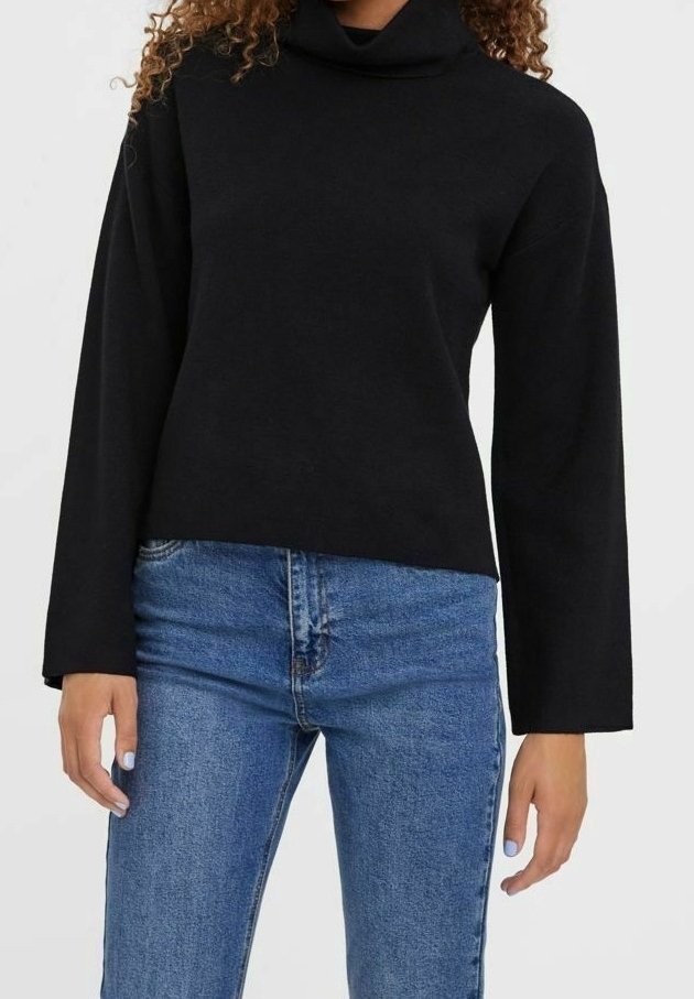 Femme portant un pull à col roulé noir à manches longues et un jean bleu en denim, debout devant un fond clair uni.