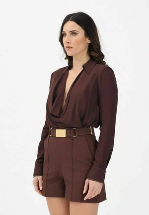 Donna con lunghi capelli scuri che indossa una blusa marrone scuro a maniche lunghe con scollo profondo e pantaloncini coordinati a vita alta con cintura con fibbia dorata.