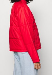 Röd puffjacka med hög krage, horisontell quiltad design och sidofickor. Materialet verkar glänsande och slätt, med en avslappnad passform.