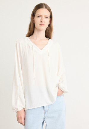 Lichtcrème blouse met lange mouwen, V-hals en trekkoorddetails. Zachte, vloeiende stof en een relaxte pasvorm. Gecombineerd met lichtblauwe spijkerbroek.