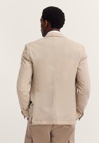 Mann mit lockigem Haar, von hinten gesehen, der ein beiges Blazerjackett und passende Cargo-Hosen vor einem einfarbigen Hintergrund trägt.