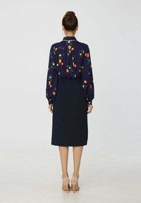 Blouse florale bleu marine avec de longues manches bouffantes et un col montant, associée à une jupe crayon noire. Des chaussures à talons beiges complètent la tenue.