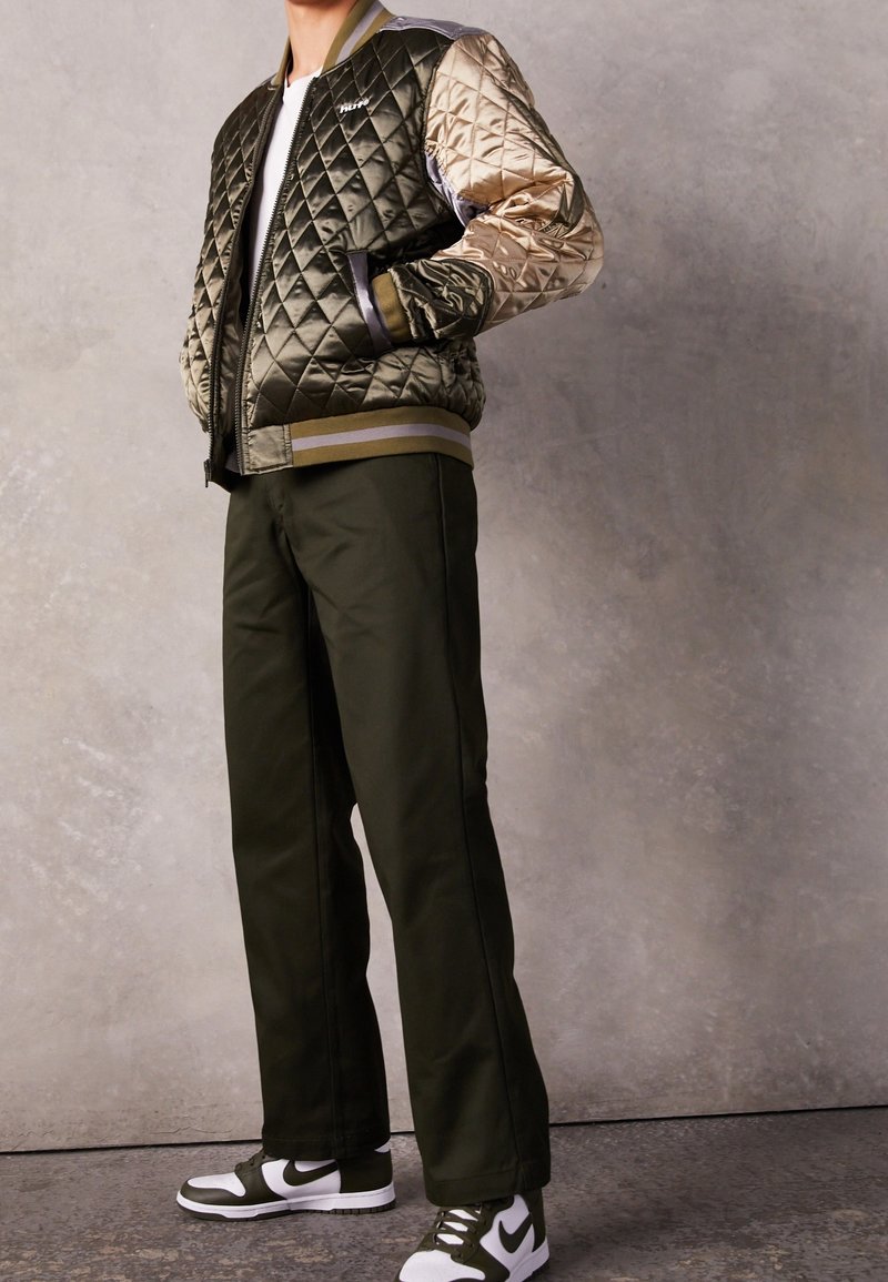 Quilted bomber jacket in olive green and beige, with ribbed cuffs and hem. Paired with olive green pants and black-and-white sneakers.