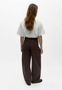 Femme aux longs cheveux bruns vue de dos, portant un t-shirt blanc ample, un pantalon large taille haute brun foncé et des chaussures noires.