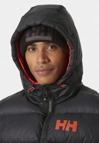 Giubbotto imbottito nero con fodera arancione, collo alto, chiusura con zip e cordini a contrasto arancioni; indossato con un berretto lavorato a maglia nero.
