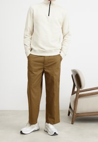 Světle béžový pulovr s krátkým zipem, hnědé kalhoty a bílé tenisky se šedými akcenty. Hladká textura, volný střih a viditelné boční kapsy.