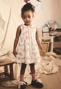 Vestito bianco senza maniche con stampe di animali colorate, bordo rosso e scollo con colletto. Scarpe marroni con fiocchi rosa e calzini rosa chiaro.