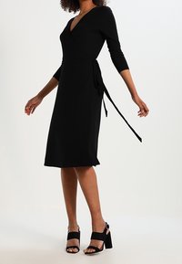 Svart ribbad omslagsklänning med V-ringning, trekvartsärmar och ett bälte i midjan. Matchas med svarta klack-sandaler med blockklack.