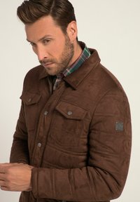 Brown suede jacket with a collared neckline, two chest pockets, silver buttons, and a textured finish, worn over a plaid shirt.