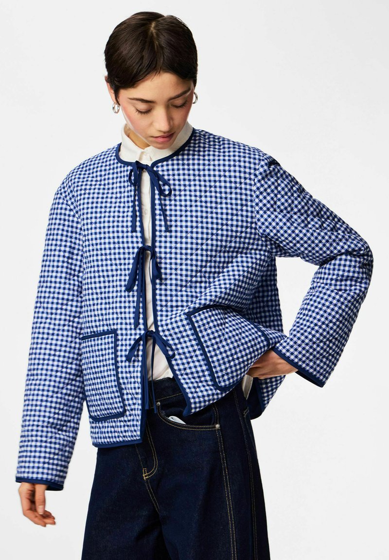 Kvinde iført blå og hvide ternede, quiltede jakke med bindebånd og lappe-lommer, kombineret med mørke denim jeans.