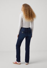 Tamsiai mėlynos platėjančios džinsai su aukšta liemene, turi dvi kišenes gale ir Lois logotipo ženklelį, derinami su balta ribuota ilgomis rankovėmis palaidine.