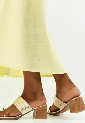 Person trägt einen hellgelben Midirock und goldene geflochtene Sandalen mit Blockabsatz, offenen Zehen und zum Hineinschlüpfen.