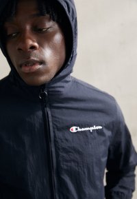 Giacca windbreaker blu marina con cappuccio, chiusura a zip e tessuto testurizzato, con il logo bianco "Champion" sul petto.
