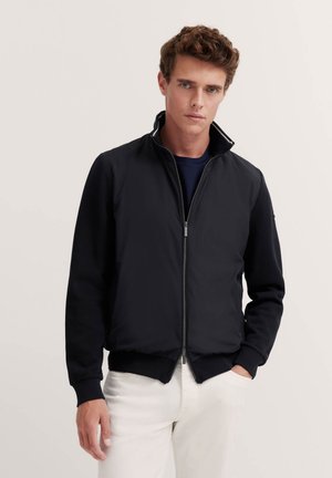 Schwarze Zip-Jacke mit kontrastierenden, strukturierten Ärmeln und Kragen, mit lockerer Passform und gerippten Bündchen. Kombiniert mit hellen Hosen.