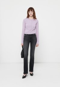 Mujer con un cárdigan lila abotonado, jeans negros de cintura alta, tacones negros, sosteniendo un pequeño bolso negro, de pie contra un fondo blanco.
