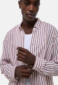 Chemise en lin rayée rose clair et blanche, boutonnée avec un col, à manches longues et coupe décontractée. Portée par-dessus un T-shirt blanc.